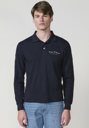 Marineblaues Langarm-Polo mit Kragen, das mit einem Brustlogo in Rot und Weiß verziert ist und aus strukturiertem Stoff besteht. Kombiniert mit hellblauen Jeans.