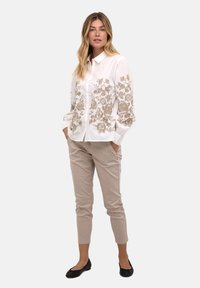 Cămașă albă cu nasturi, cu broderie florală bej pe mâneci, asortată cu pantaloni bej și balerini negri. Modelul Poză cu mâinile în buzunare.