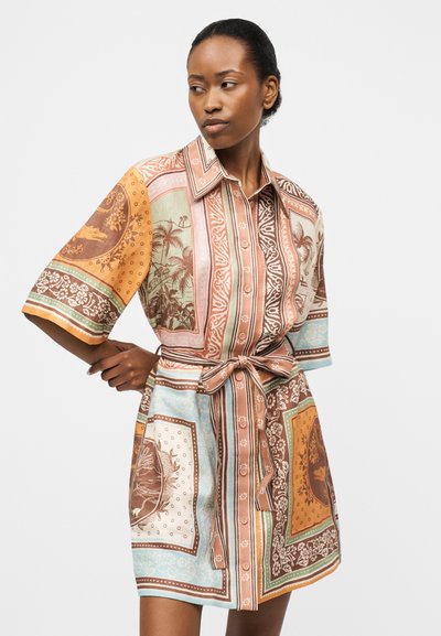 Γυναίκα που φοράει φόρεμα-πουκάμισο με σχέδιο, κουμπωτό, με μανίκια μέχρι τον αγκώνα και ζώνη στη μέση, κοιτάζοντας προς τα αριστερά της, μπροστά σε λευκό φόντο.