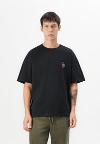 Sort bomulds T-shirt med løs pasform, korte ærmer og et lille broderet design på brystet. Enkel tekstur og rund halsudskæring.