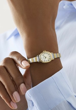 Orologio da polso con cinturino in argento e oro su un polso dalla pelle scura, indossato sopra il polsino di una camicia azzurra con bottoni.