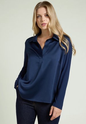 Kvinna med långt blont hår som bär en mörkblå långärmad blus med krage och mörka jeans, stående med ena handen i fickan mot en enhetlig bakgrund.