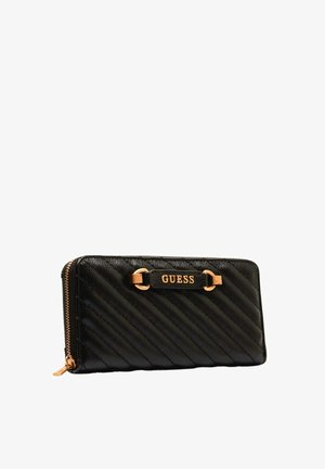 Portefeuille Guess en cuir matelassé noir avec fermeture éclair et plaque logo dorés sur le devant, vue en angle sur fond blanc.