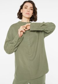 Langarmshirt in Olivgrün mit Stehkragen, Knopfleiste und hochgerollten Ärmeln. Hergestellt aus einem weichen, glatten Stoff mit lässigem Schnitt.