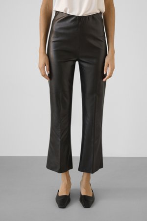 Person wearing black faux leather cropped pants with a high waist and black square-toe shoes, standing on gray floor against white background.