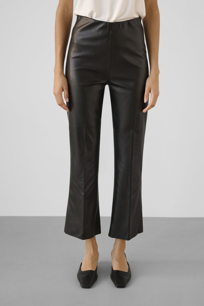 Isik, kes kannab musti kunstnahast lühendatud pükse kõrge vöökohaga ja musti ruudukujulise ninaosaga kingi, seisab hallil põrandal valge tausta ees.