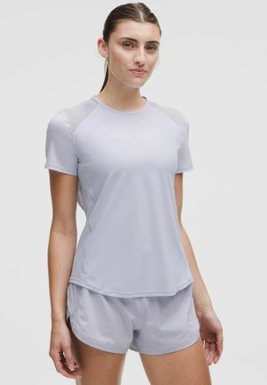 Young woman standing wearing a light purple short-sleeve athletic shirt and matching shorts against a plain background.