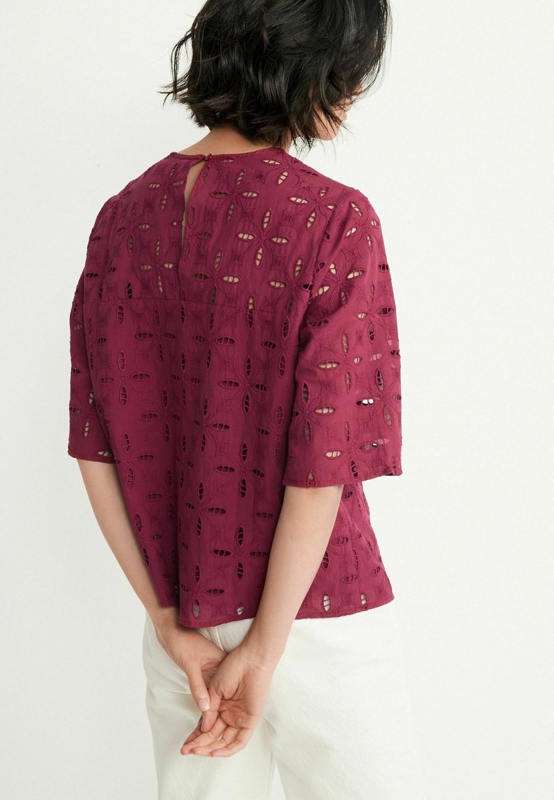 Femme aux cheveux courts et foncés portant un chemisier ajouré bordeaux à manches courtes et un pantalon blanc, tournée de dos, les mains jointes dans le dos.