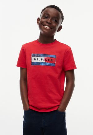 Lächelnder Junge, der ein rotes T-Shirt mit einem blau-weißen und roten Logo sowie dunkle Hosen trägt, Hände in den Taschen, vor einem einfachen weißen Hintergrund.