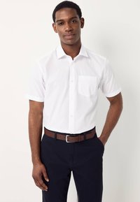 Chemise blanche à manches courtes avec boutons et poche, associée à un pantalon bleu marine foncé et une ceinture marron. Tissu lisse avec une coupe ajustée.