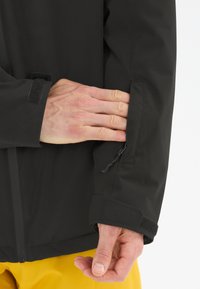 Schwarze wasserdichte Jacke mit Nahaufnahme des Bündchens, das mit Klettverschluss und Reißverschlusstasche ausgestattet ist. Glatte Textur und taillierte Passform.