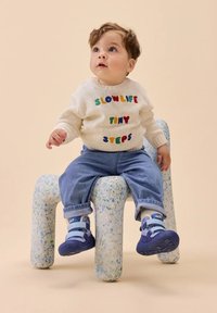 Weißer Strickpullover mit mehrfarbigem Text, blaue Jeanshose und blaue Schuhe mit Klettverschluss. Kind sitzt auf einem gesprenkelten, abstrakt geformten Hocker.