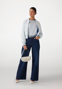 Giacca trapuntata blu chiaro, t-shirt grigia con ricamo, jeans in denim scuro a gamba larga, scarpe bianche con tacco alto e una piccola borsa beige.