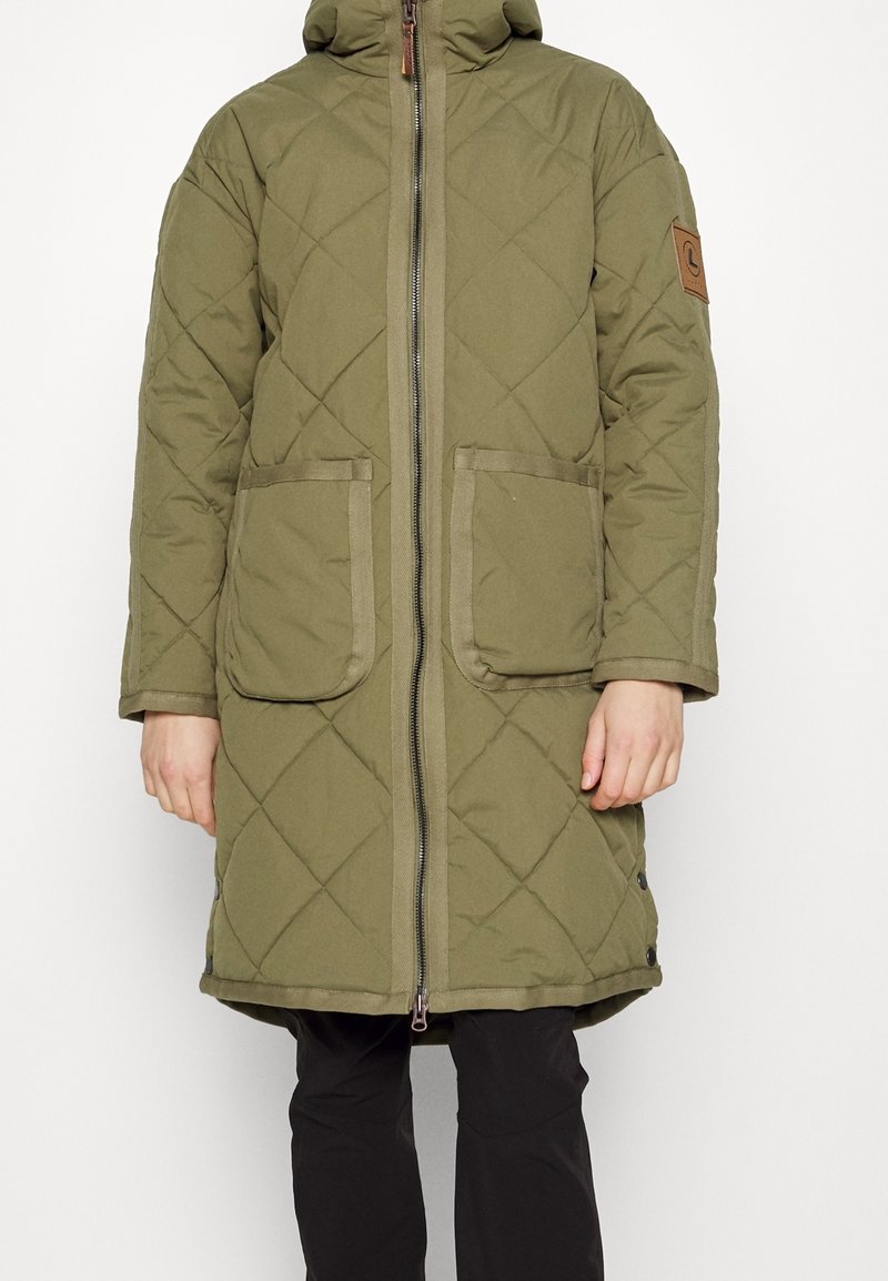 Long manteau matelassé vert olive avec fermeture éclair à l'avant, deux grandes poches avant, capuche et une écusson marron sur le haut du bras, porté avec un pantalon noir.