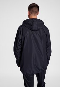 Chaqueta con capucha negra, hecha de material ligero, que presenta un corte relajado, puños elásticos y un dobladillo con cordón.