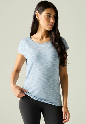 Mujer con cabello largo y oscuro, que lleva una camiseta azul clara de manga corta y leggings negros, ajustando el dobladillo de su camiseta.