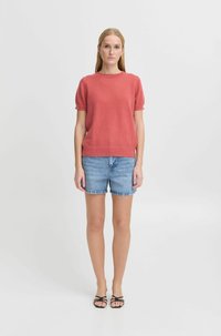 Korallfärgad t-shirt med korta ärmar i en mjuk textur, kombinerad med ljusblå denimshorts. Modellen bär svarta sandaler med remmar.