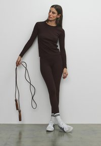 Bruine top met lange mouwen en bijpassende leggings van een glad, rekbaar stof. Het model houdt een bruune springtouw met houten handgrepen vast en draagt witte sneakers.