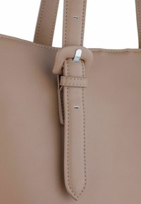 Close-up van een beige leren tasriem met een rechthoekige, gewatteerde gesp en een zilveren pin, waarbij de gedetailleerde stiksels en de verstelgaten zichtbaar zijn.