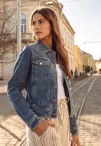 Jonge vrouw in spijkerjasje en gestreepte broek staat op een geplaveide straat vlak bij tramrails in een zonnige stedelijke omgeving.