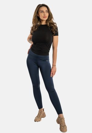 Maglietta nera a maniche corte abbinata a leggings fitness blu navy, con una consistenza liscia e un discreto dettaglio del logo vicino all'elastico. Calzature sono sneakers color beige.