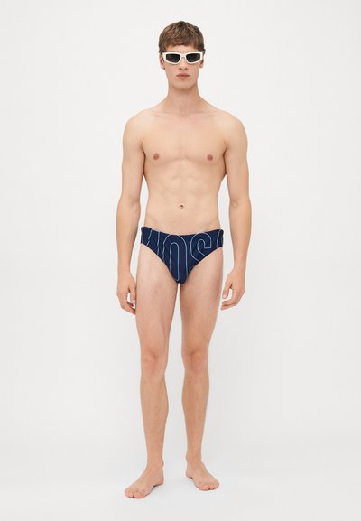 Junger Mann steht barfuß und trägt dunkelblaue Badehose mit hellblauen vertikalen Streifen und weiße Sonnenbrille vor einfarbigem Hintergrund.