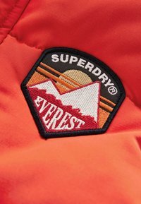 Tissu rouge avec un écusson brodé bordé de noir représentant des montagnes, un soleil et le texte "SUPERDRY EVEREST" en blanc et orange.