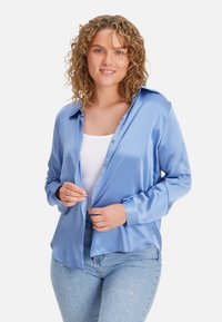 Light blue satin shirt with long sleeves, button closure, and a collar. Worn open over a white top and paired with light denim jeans.