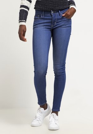 Mujer con vaqueros ajustados azules, zapatillas blancas y un suéter de rayas blancas y negras, de pie frente a un fondo claro y liso.