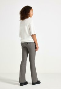 Nainen, jolla on kiharat hiukset, pukeutuneena valkoiseen lyhythihaiseen neuleeseen, harmaisiin slim fit -housuihin ja mustiin platform-kenkiin, seisoo valkoista taustaa vasten.