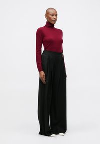 Burgundi turtleneck pulóver, laza fekete nadrág pliszével és fehér hegyes orrú cipők. Simább textúrák, testre szabott dizájn és diszkrét részletek.