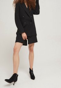 Blazer negro de doble botonadura con solapas con muesca, combinado con pantalones cortos negros a juego. Botines de tacón negro añaden contraste y textura.