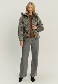 Prešívaná sivá puffer bunda s kapucňou, so geometrickým vzorom a elastickými manžetami, skombinovaná s vysokými sivými džínsami a čiernymi špicatými topánkami.