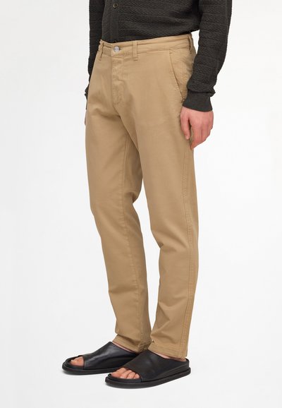 Homme portant un pantalon beige et des sandales noires ouvertes à l'avant, de type slip-on, avec un haut texturé sombre à manches longues, debout devant un fond blanc.