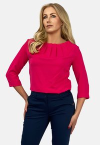 Fuksjowa bluzka z plisowanym dekoltem i rękawami 3/4, połączona z ciemnoniebieskimi spodniami o wąskim kroju, bez widocznych wzorów czy dodatków.