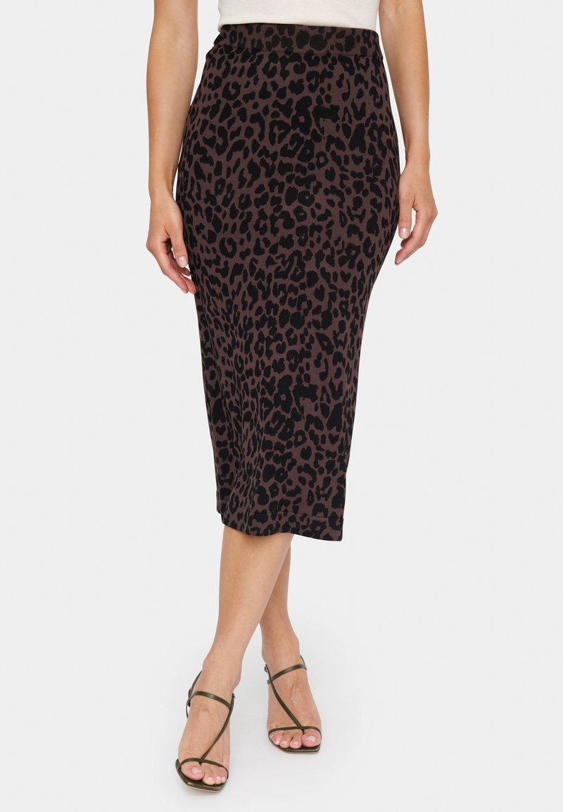 Femme portant une jupe crayon midi marron à imprimé léopard noir, associée à des sandales à talons hauts noires à lanières et un haut blanc.