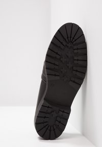 Botte à semelle en caoutchouc noir positionnée debout montrant des crampons profonds avec des motifs en croix et en ligne sur un fond d’angle blanc.