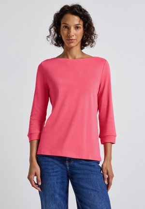 Vrouw met krullend donker haar draagt een koraalroze top met driekwartmouwen en een blauwe spijkerbroek, terwijl ze tegen een effen lichte achtergrond staat.