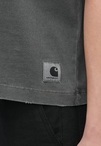 Pelēks kokvilnas T-kreklis ar nedaudz nolietotu apakš malu, ar melnbaltu kvadrātveida Carhartt logo birku apakšējā kreisajā stūrī.