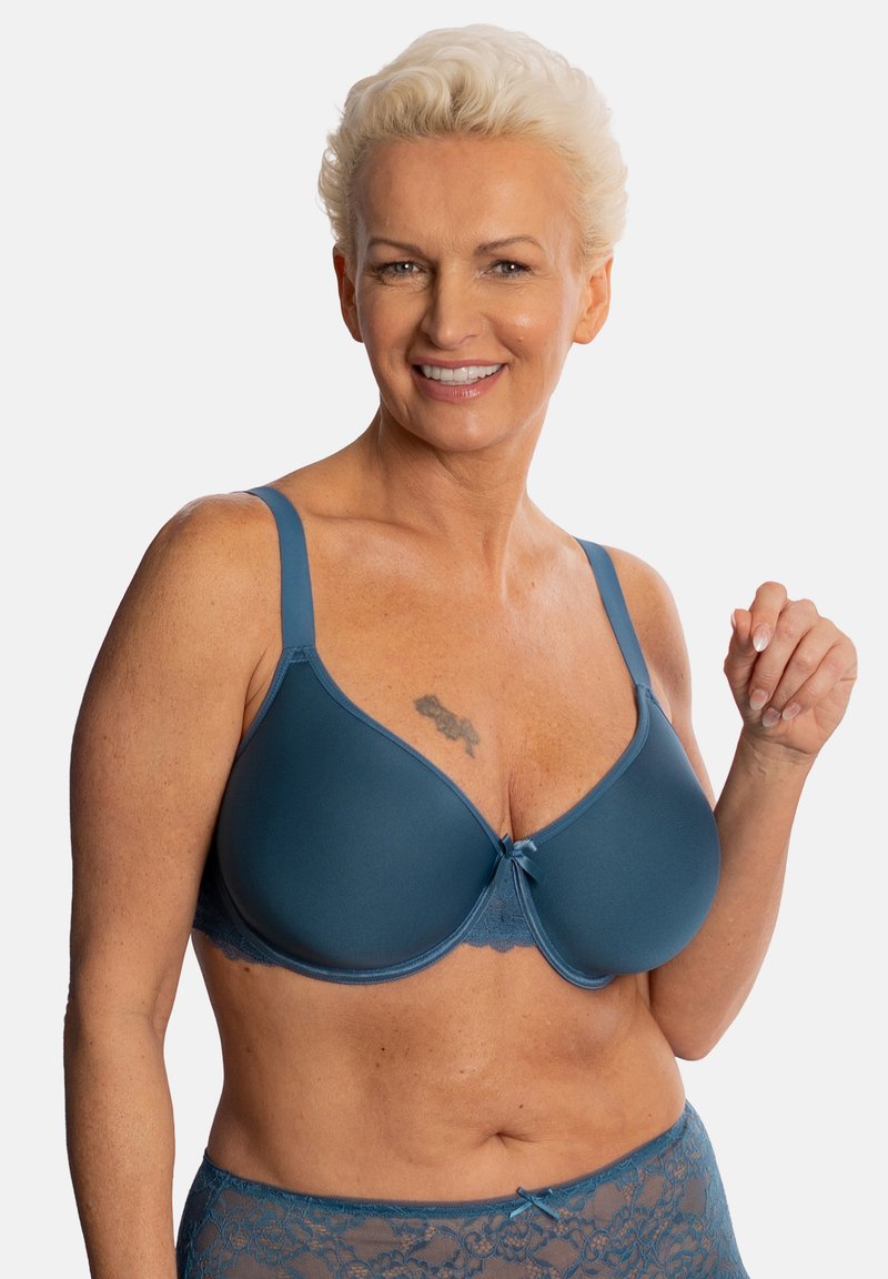 Mature woman with short blonde hair smiling, wearing teal smooth cup bra and matching lace underwear, with visible chest tattoo.