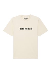 T-shirt crema con maniche corte e scollo rotondo. Presenta un tratto di testo nero audace che recita "FISSA SE TI PIACCIO" sul davanti.