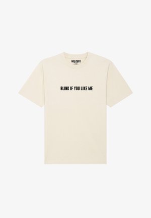 T-shirt crème à manches courtes et col rond. Présente un texte en gras noir affichant "CLIGNE SI TU M'AIMES" sur le devant.