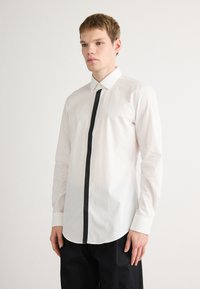 Witte overhemd met knopen, voorzien van een contrasterende zwarte verticale streep aan de voorkant. Heeft een klassieke kraag en lange mouwen.