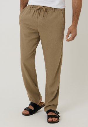 Mand iført beige bukser med snøre og sorte sandaler med to stropper, stående med den ene hånd afslappet ned langs siden.