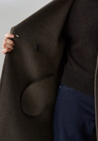 Een hand houdt een donkergrijze wollen jas open, zichtbaar is een zwarte knoop en een binnenzak, terwijl een persoon blauwe jeans en een donkere trui draagt.