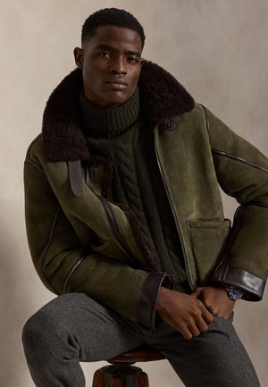 Jeune homme portant une veste en shearling vert foncé, un col roulé en maille verte, un pantalon gris et une montre bleue, assis sur un tabouret en bois devant un fond neutre.