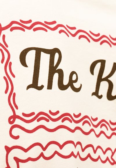 Καφέ κείμενο με καλλιγραφικά γράμματα "The K" πλαισιωμένο από διακοσμητικές κόκκινες κυματιστές γραμμές σε λευκό υφασμάτινο φόντο.