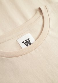 Beige bomulds t-shirt med ribbet rund halsudskæring. Har et vævet mærke med bogstavet "W" på halsen. Blød tekstur og glat finish.