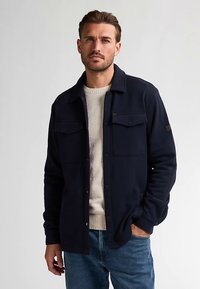 Donkerblauw overhemdjack gemaakt van zacht stof, met twee borstzakken, knoopsluiting en een relaxte pasvorm gedragen over een crèmekleurige trui.