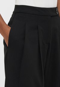 Μαύρο παντελόνι σε στενή γραμμή με ψηλή μέση, δύο πλαϊνές τσέπες, πιέτες στο μπροστινό μέρος και διπλό κουμπί στη μέση.